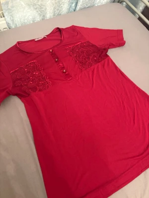 Röd topp med spetsdetaljer Rozalinda - Röd topp från Rozalinda med korta ärmar, rund halsringning och dekorativa spetsdetaljer framtill. Toppen har knappar vid bröstet och är tillverkad i mjukt material som känns skönt mot huden. Perfekt för dig som gillar färg och fina detaljer. kan även vara sovplagg