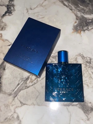 Versace Eros Eau de Toilette 100ml - Versace Eros Eau de Toilette i en elegant, fyrkantig blå glasflaska med detaljerat mönster och Versace-logga. Flaskan har ett matchande blått lock och kommer i en lyxig blå kartong med diskret mönster och silvertext. En ikonisk doft för dig som gillar exklusiva parfymer.