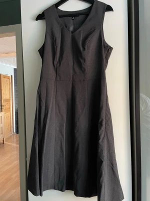 Mörkgrå ärmlös klänning med v-ringning - Snygg mörkgrå klänning med v-ringad hals och ärmlös design. Klänningen har en klassisk, lätt utställd form och är fodrad på insidan för extra komfort. Perfekt för dig som gillar stilrena och enkla plagg.