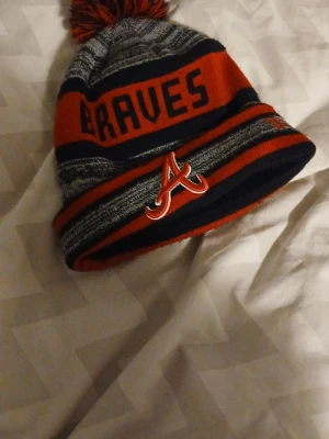 Braves mössa med tofs från New Era - Säljer en snygg stickad mössa från New Era med Braves-tryck och broderad logga framtill. Mössan har röd, marinblå och grå färg, samt en stor tofs på toppen. Perfekt för dig som gillar sportig stil och vill hålla dig varm.