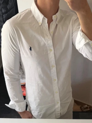Vit skjorta från Ralph Lauren - Klassisk vit skjorta från Ralph Lauren med button-down krage och broderad marinblå logga på bröstet. Skjortan har långa ärmar, knäppning framtill och är tillverkad i bomull. Perfekt för en stilren och tidlös look. Lite missfärgad och har en ganska stor lagning vid armbågen som man ser på sista bilden, därav priset. 