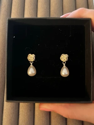 Droppformade pärlörhängen i silver - Säljer ett par eleganta örhängen med droppformade vita pärlor och detaljer i silverfärgad metall. Örhängena har en liten blomliknande dekoration upptill och klassisk stiftbaksida. Perfekta för dig som gillar stilrena accessoarer med en twist.