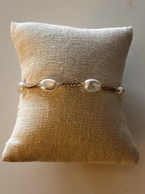 Guldarmband med vita pärlor - Snyggt armband med guldfärgad kedja och ovala vita pärlor. Kedjan har en enkel karbinhake och pärlorna är jämnt fördelade längs hela armbandet. Perfekt accessoar för att lyfta din stil.