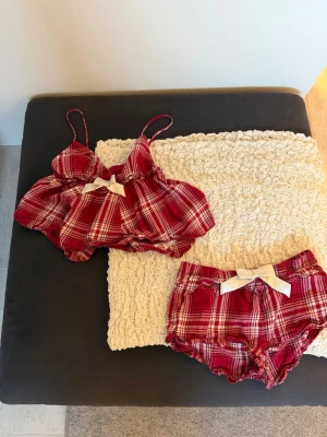 Rött rutigt pyjamasset med rosett - Supersött pyjamasset i röd- och vitrutigt med linne och shorts. Linnet har smala axelband och en dekorativ vit rosett framtill. Shortsen har matchande rosett och volangkant. Perfekt för mysiga kvällar! Materialet känns mjukt och luftigt.