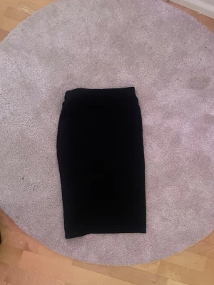 Svart pennkjol med slits - En svart pennkjol i klassisk modell med en liten slits framtill. Kjolen är midilång och har en figurnära passform som framhäver siluetten. Perfekt att styla med en crop top eller en oversized hoodie för en cool look.
