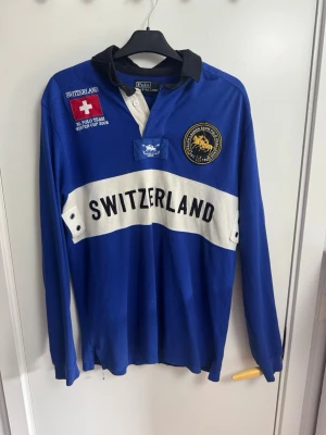 Blå långärmad poloshirt Switzerland - Blå långärmad poloshirt från Polo Team Ralph Lauren med vit panel och texten SWITZERLAND framtill. Märkta detaljer och broderade patches på bröstet samt stor romersk siffra IV på ryggen. Klassisk krage i svart och vita knappar. Perfekt för dig som gillar sportig stil.