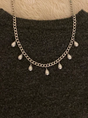 Silverfärgat halsband med droppformade pärlor - Stilrent halsband i silverfärgad rostfritt med sju droppformade vita pärlor som hänger längs kedjan. Kedjan är grov och ger ett trendigt intryck, perfekt för att lyfta din outfit med en elegant touch.