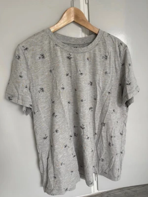 Grå t-shirt med palmmönster Large - Snygg grå t-shirt med små svarta palmtryck över hela plagget. T-shirten har rund halsringning och korta ärmar, perfekt för en avslappnad stil. Tillverkad i mjuk bomull som känns skön mot huden.