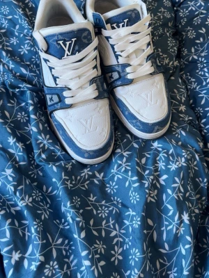 Louis Vuitton blåvita sneakers - Snygga Louis Vuitton sneakers i vitt och blått med klassiskt LV-märke på tungan och sidan. Skorna har vita skosnören, blå detaljer i denim-look och präglade LV-loggor på tån. Låg modell med robust sula och exklusiv känsla. Perfekta för dig som gillar streetstyle och designer. Storlek 43