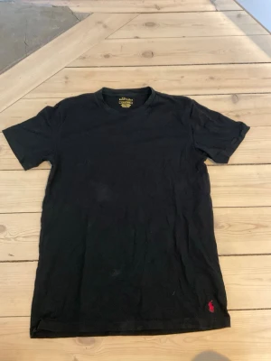 Svart t-shirt från Ralph Lauren - Klassisk svart t-shirt från Ralph Lauren med röd broderad logga nertill. T-shirten har rund halsringning och korta ärmar. Tillverkad i mjuk bomull för en skön känsla och enkel stil.