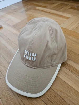 Beige keps från Miu Miu - Snygg beige keps från Miu Miu med vit broderad logga framtill. Kepsen har böjd skärm och vit kant runt om, vilket ger en clean och modern look. Tillverkad i bomullstyg som känns både bekvämt och stilrent.