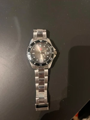 Silverfärgad Swiss Military klocka - Stilren klocka från Swiss Military med silverfärgad länk i metall och svart urtavla. Klockan har tydliga siffror på vridringen, datumvisning och klassisk design med Swiss Military-loggan på urtavlan. Perfekt accessoar för dig som gillar en sportig men ändå clean look.