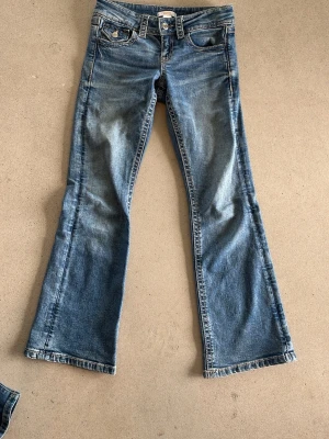 Blå bootcut jeans från Gina Tricot - Säljer ett par blå bootcut jeans från Gina Tricot med klassisk femficksmodell och snygga kontrastsömmar. Jeansen har en lätt utsvängd passform nedtill och är tillverkade i slitstark denim. Perfekta för en avslappnad och trendig look.