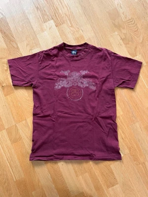 Rare made in usa stüssy T-shirt  - Säljer en vinröd t-shirt från Stüssy med korta ärmar och ett tryck och text på bröstet. T-shirten har en klassisk passform och är gjord i mjuk bomull. Tröja är i mycket fint skick med tanke på hur gammal den är, bara att det finns lite små hål på den högra ärmen. Hör av er om ni har frågor!!