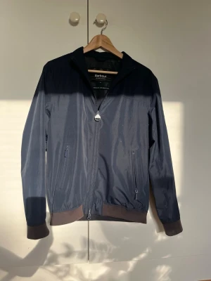 Marinblå vindjacka från Barbour - Snygg marinblå vindjacka från Barbour med dragkedja framtill och två fickor med dragkedja. Jackan har ribbade muddar och nederkant i mörkare ton. Perfekt för blåsiga dagar och enkel att matcha med olika outfits.