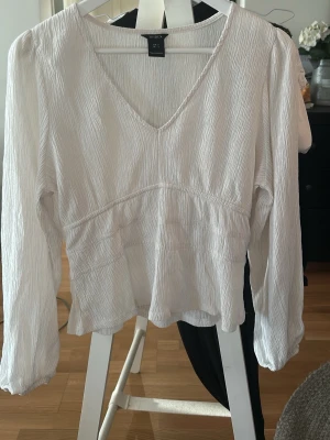 Söt top från LINDEX - Stilren vit blus från LINDEX med v-ringning och långärmad design. Tillverkad i ett lätt, krinklat tyg som ger en avslappnad men ändå chic look. Perfekt att matcha med både jeans och kjol för en trendig outfit.