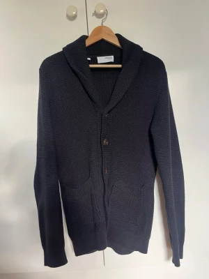 Mörkblå kofta med sjalkrage - Stilren mörkblå kofta från Selected Homme med sjalkrage och knäppning framtill. Koftan har två framfickor och är stickad i ett grovt material som ger en avslappnad look. Perfekt att slänga över en t-shirt för extra lager.