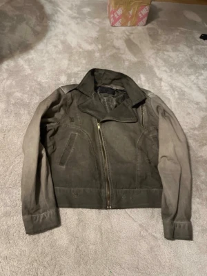 Grå vintage skinnjacka med dragkedja - Säljer en grå skinnjacka med klassisk krage och sned dragkedja. Jackan har två snedställda fickor framtill och detaljerade sömmar. Passar dig som vill ha en cool och tidlös look. Perfekt att matcha med jeans och t-shirt.
