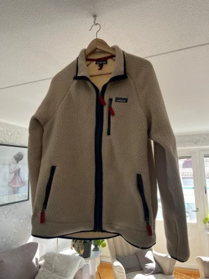 Beige fleecejacka från Patagonia - Mysig beige fleecejacka från Patagonia med hel dragkedja framtill, mörkblå detaljer och röda dragkedjor. Jackan har två sidofickor och en bröstficka med dragkedja. Perfekt för lager-på-lager och outdoor-häng.