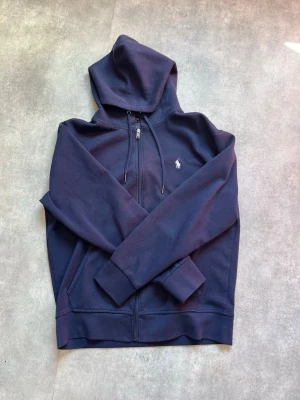 Mörkblå hoodie från Polo Ralph Lauren - Mörkblå hoodie med dragkedja från Polo Ralph Lauren. Klassisk design med huva och dragsko, två fickor framtill och den ikoniska broderade loggan på bröstet. Perfekt för en avslappnad och stilren look.