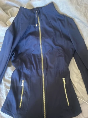 Marinblå träningsjacka från Lululemon - Snygg marinblå träningsjacka från Lululemon med figurnära passform. Jackan har guldfärgade dragkedjor framtill och på fickorna samt detaljerade sömmar för en sportig look. Tillverkad i stretchigt funktionsmaterial som andas. Jag har haft den väldigt länge och använt 1 gång, säljer då den Inte används😊🥰