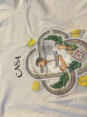 Vit Casa Blanca t-shirt med tennistryck - Vit t-shirt från Casa Blanca med färgglatt tryck av en tennisspelare, tennisbollar och gröna blad. T-shirten har rund hals och korta ärmar. Perfekt för dig som gillar sportig och unik stil.