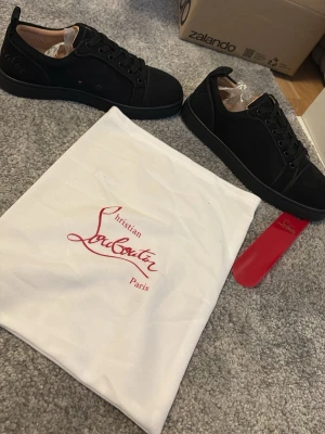Svarta sneakers från Christian Louboutin - Stilrena svarta sneakers från Christian Louboutin med klassisk röd sula och svarta skosnören. Skorna har en låg siluett och är tillverkade i ett matt material med diskreta detaljer. Perfekta för dig som vill ha en lyxig och trendig look.