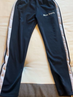 Palm Angels svarta mjukisbyxor - Palm Angels mjukisbyxor i svart med vita sidoränder och logga på vänster lår. Byxorna har en avslappnad passform, resår i midjan och sidofickor. Perfekta för en chill och sportig look.