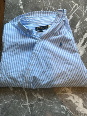 Blåvit randig skjorta från Polo Ralph Lauren - Snygg blå och vit randig skjorta från Polo Ralph Lauren i klassisk passform. Skjortan har button down-krage, broderad logga på bröstet och är tillverkad i tunn bomull. Perfekt för en fräsch och stilren look.