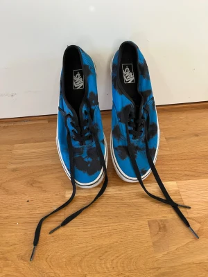 Blå tie-dye Vans sneakers - Säljer ett par blå Vans sneakers med svart tie-dye mönster och svarta skosnören. Klassisk låg modell med vit sula och den ikoniska gummisulan undertill. Perfekta för dig som gillar streetwear och vill sticka ut lite extra.