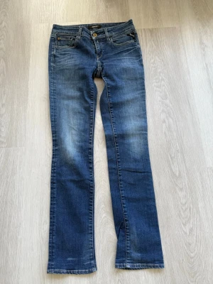 Blå bootcut jeans från Replay - Snygga blå bootcut jeans från Replay med klassisk femficksdesign och lätt slitna detaljer. Jeansen har normal midja, gylf med knapp och dragkedja samt logotyp på fickan. Perfekta för en avslappnad och trendig look.