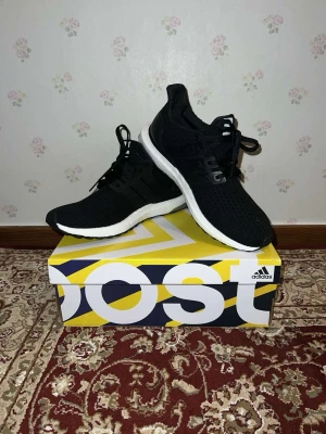 Svarta Adidas Ultra Boost sneakers - Säljer ett par svarta Adidas Ultra Boost sneakers med stickad ovandel och vita Boost-sulor. Skorna har klassiska tre ränder på sidan, svarta snören och en bekväm, sportig design. Perfekta för dig som gillar stilrena och sköna sneakers.