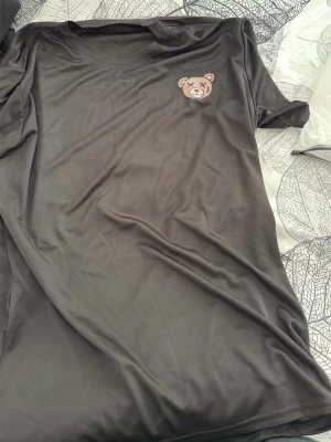 Svart t-shirt med björnmärke - Snygg svart t-shirt med ett litet tryck av en björn med korsade ögon på bröstet. T-shirten har rund hals och korta ärmar. Materialet känns mjukt och lätt, perfekt för en avslappnad stil.