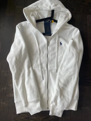 Vit hoodie från Polo Ralph Lauren - Helt ny och oanvänd vit zip hoodie från Polo Ralph Lauren med broderad mörkblå logga på bröstet. Storlek M men liten i storleken, passar även S. Stilren och perfekt till vardag