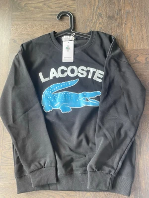 Svart sweatshirt från Lacoste M - Helt ny och oanvänd svart Lacoste sweatshirt med stort tryck framtill. Storlek M men liten i storleken, passar även S. Stilren och skön tröja som passar till allt
