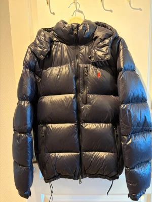 Svart pufferjacka från Polo Ralph Lauren - Snygg svart pufferjacka från Polo Ralph Lauren med röd broderad logga på bröstet. Jackan har hög krage, dragkedja framtill, flera fickor med dragkedja och justerbar nederkant. Perfekt för kalla vinterdagar tack vare det vadderade materialet.
