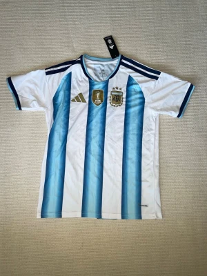 Argentina fotbollströja till VM - Säljer en Argentina fotbollströja från Adidas med klassiska blå och vita ränder, guldiga FIFA- och AFA-emblem på bröstet samt mörkblå detaljer på axlar och ärmslut. Tillverkad i lätt och ventilerande material, perfekt för träning eller match.
