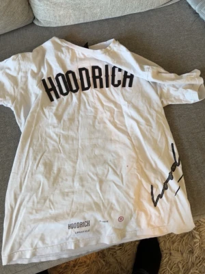 Vit HOODRICH t-shirt med tryck - Vit t-shirt från HOODRICH med svart tryck på ryggen och framtill. Klassisk passform med korta ärmar och rund hals. Stort HOODRICH-tryck på ryggen och mindre tryck nertill. Perfekt för en streetwear-look.