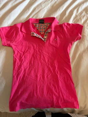 Rosa pikétopp från Tommy Hilfiger - Säljer en snygg rosa pikétopp från Tommy Hilfiger Denim med klassisk krage och korta ärmar. Insidan av kragen har ett rutigt mönster i rött, blått och vitt som ger en cool detalj. Perfekt för dig som gillar stilrena och färgglada plagg. Vet inte om den är äkta 