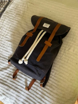 Ryggsäck från Sandqvist - Ryggsäck från Sandqvist med bruna läderdetaljer och vita snören. Justerbara axelremmar och praktiskt innerfack. Tillverkad i slitstark canvas och läder, perfekt för skolan eller stan.