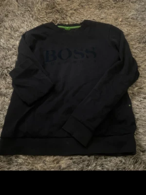 Svart sweatshirt från BOSS - Svart sweatshirt från BOSS med diskret logga framtill. Tröjan har rund halsringning, långa ärmar och ribbade muddar. Slim fit-modell med gröna detaljer på insidan av kragen. Perfekt för en stilren och avslappnad look. Stl L sitter som M