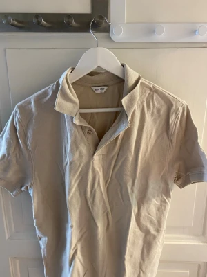 Beige pikétröja från East West - Säljer en stilren beige pikétröja från East West i storlek M. Tröjan har korta ärmar, klassisk krage och knappar vid halsen. Perfekt till jeans eller shorts för en clean look.