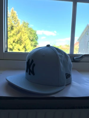 Ljusblå New Era NY keps - Säljer en ljusblå snapback keps från New Era med klassisk New York Yankees-logga i svart framtill. Kepsen har platt skärm och justerbar baksida för perfekt passform. Tillverkad i bomullstyg och har broderade ventilationshål för extra komfort. Storleken är justerbar och är Medium/Large 
