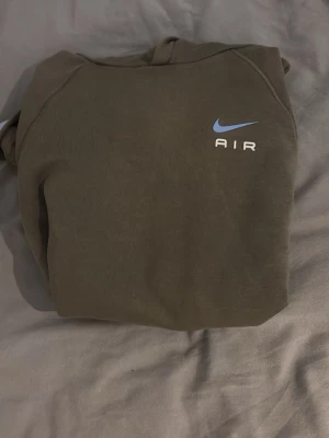 Nike Air hoodie i olivgrönt - Snygg olivgrön hoodie från Nike Air med blå logga och text på bröstet. Klassisk känguruficka framtill och huva. Perfekt för en avslappnad streetwear-look. Mjuk bomullsblandning som känns skön mot huden.