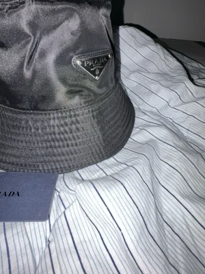 Svart bucket hat från Prada - Snygg svart bucket hat från Prada i glansigt nylonmaterial. Klassisk form med bred, nedåtlutande brätte och ikonisk metalltriangel med logga framtill. Perfekt accessoar för en trendig och avslappnad stil.