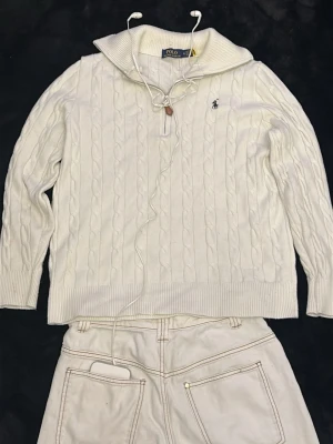 Vit kabelstickad tröja Polo Ralph Lauren - Kabelstickad vit tröja från Polo Ralph Lauren med halv dragkedja och ribbad krage. Tröjan har broderad logga på bröstet och är gjord i mjukt material. Perfekt för en clean och klassisk stil.