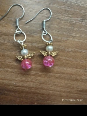 Örhängen med ängel och pärlor - Säljer ett par unika örhängen med ängelmotiv. De har vita pärlor som huvud, rosa glittrande pärlor som kropp och små guldfärgade vingar. Krokarna är i silverfärgad metall. Perfekt accessoar för dig som gillar söta detaljer.