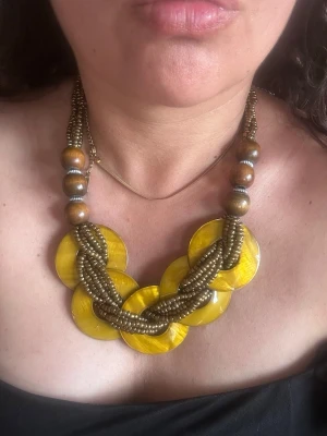 Gult halsband med pärlor och trädetaljer - Unikt halsband med stora runda gula plattor, flätade bronsfärgade pärlor och träkulor. Halsbandet har en bohemisk vibe och är perfekt för att ge din outfit en färgklick. Materialen är plast, trä och metallpärlor. Ingen synlig märkning.