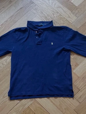 Ralph Lauren långärmade piké - Klassisk mörkblå henley från Polo Ralph Lauren med lång ärm och liten broderad logga på bröstet. Skjortan har knappar vid halsen och en mjuk bomullskänsla. Perfekt för en avslappnad men ändå stilren look.