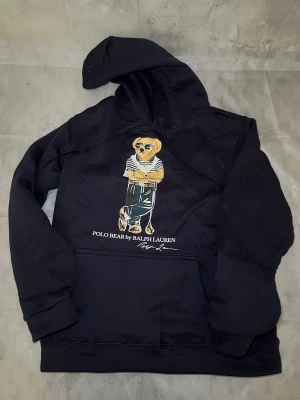 Svart hoodie Polo Bear Ralph Lauren - Mörk blå hoodie från Ralph Lauren med Polo Bear-tryck framtill. Klassisk huva och känguruficka. Björnen på framsidan har solglasögon och randig tröja, vilket ger en cool och avslappnad vibe. Perfekt för dig som gillar streetwear och ikoniska prints.